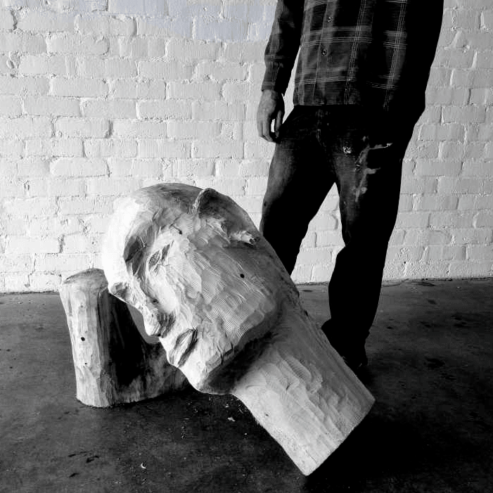artist standing next to wooden sculpture of a head leaning against a wooden plinth, brick wall, and floor